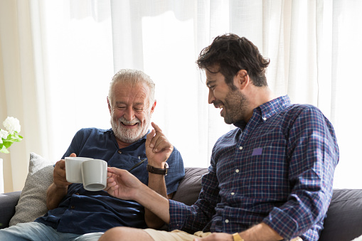 Senior father with adult son talking and drinking coffee while sitting on sofa at home with happy