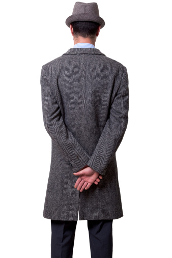 A person dressed in a gray overcoat and a gray hat standing with his back to the camera, holding his right hand over his left one. .Isolated on white background.