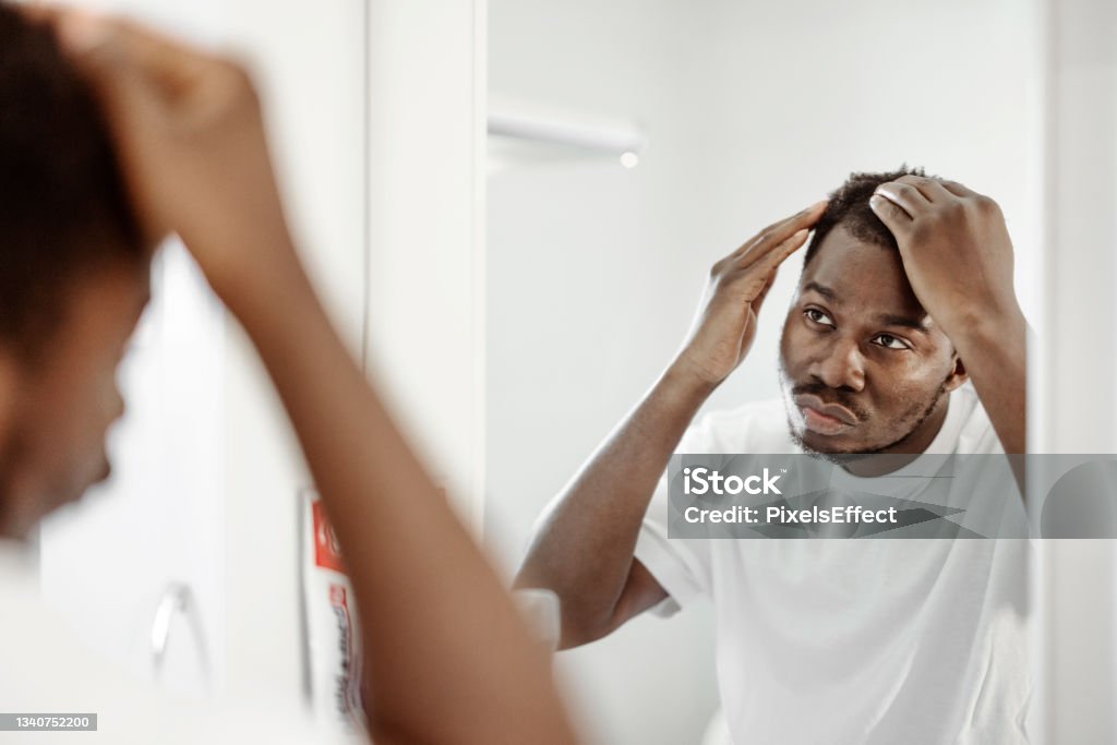 Man Worrying About Hair Loss Hair loss man looking in bathroom mirror touching his hair or checking for hair loss problem. Male problem of losing hairs Alopecia Stock Photo