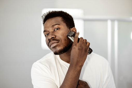 Young man shaving with electric razor and looking at camera