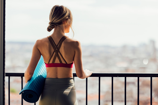 Back view of unrecognizable fit young woman in sportswear standing on balcony and looking at city