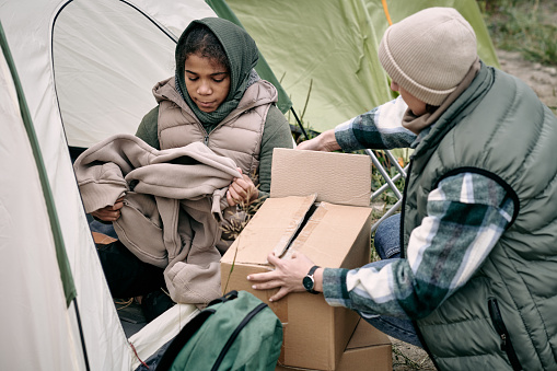 Middle-eastern refugee family in warm clothes unpacking boxes while getting settled into tent after escape from town