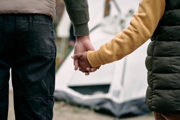 Holding Hand of Child Against Migrant Camp Rear view of unrecognizable parent of elder brother holding hand of child against tent in migrant camp refugee photos stock pictures, royalty-free photos & images