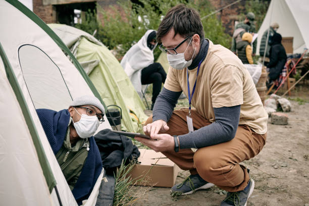 Social Worker Talking To Refugee Young social worker with badge crouching at tent and making notes in tablet while talking to refugee homelessness stock pictures, royalty-free photos & images