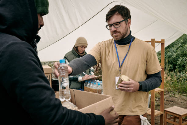 Volunteer Giving Water To Homeless Man Young bearded volunteer in glasses holding tin can and giving bottle of water to homeless man with box water crisis stock pictures, royalty-free photos & images