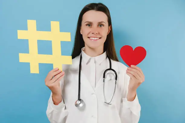Photo of Portrait of friendly young woman doctor with toothy smile have good mood holding large big yellow hashtag sign and little red heart, wears coat, posing isolated over blue color background in studio