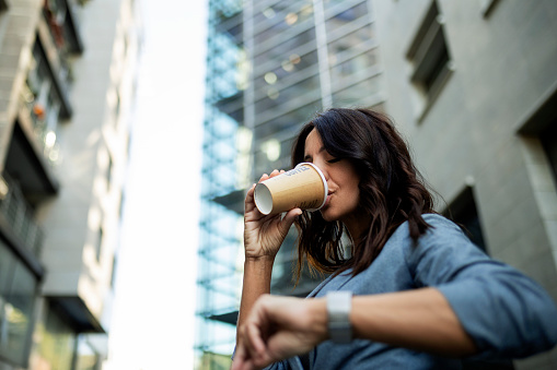 Young business woman looking at her wristwatch outside office building drinking coffee