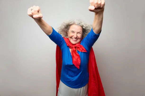 Mature woman,57 years old,wearing a blue shirt and a red cape made from a piece of fabric, her arms are raised, posing as superwoman, hair is blown away, with the help of a fan ,as if flying. The woman has beautiful long, grey and curly hair