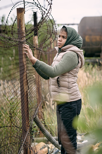 Sorrowful middle-eastern girl in headscarf and vest lost her parents during hostilities standing at barbed wire