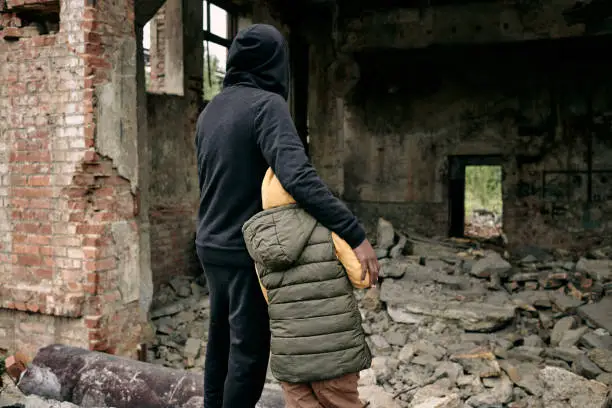 Rear view of father embracing child and looking at ruins of house after hostilities