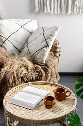Concept of recreation, relaxation. Weekend at cozy house. Wicker coffee table with open book and wooden cup with beverage standing near rattan armchair. Living room interior at boho chic style
