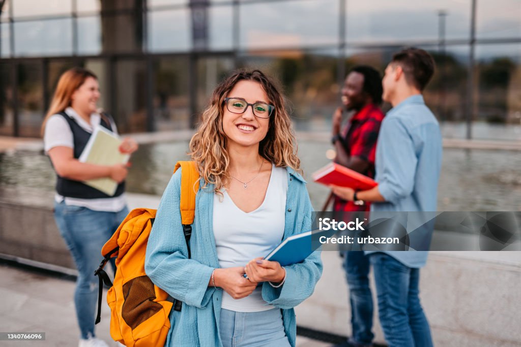 Beautiful smiling female college student Portrait of a smiling female university student standing on campus with friends in the background. University Student Stock Photo