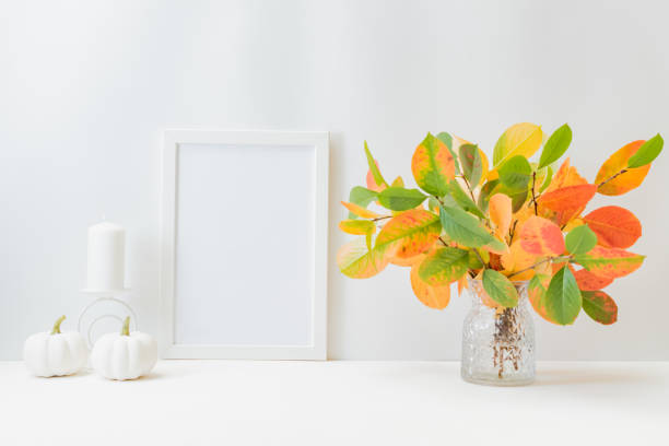 istock Home interior with decor elements. Mockup with a white frame, colorful autumn leaves in a vase on a light background 1340766181