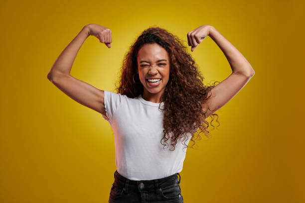 portrait en studio d’une jeune femme fléchissant ses biceps sur un fond jaune - flexing muscles photos et images de collection