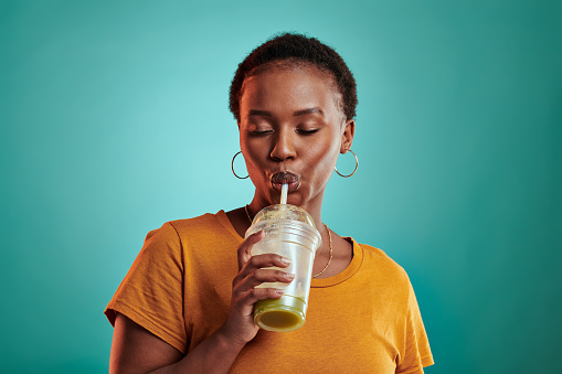 Shot of a young woman drinking a green juice while standing against a turquoise background