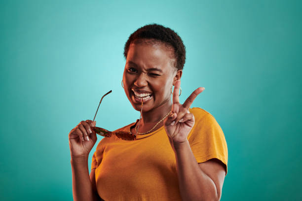 Shot of a woman holding sunglasses and showing the peace sign while standing against a turquoise background stock photo