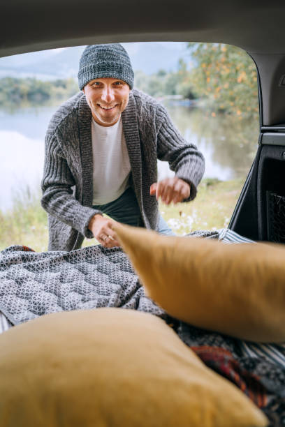 istock Sincerely smiling middle-aged man dressed in warm knitted clothes preparing a sleeping place in cozy car trunk on beautiful mountain lake bank. Warm early autumn auto camper traveling concept image. 1340785446