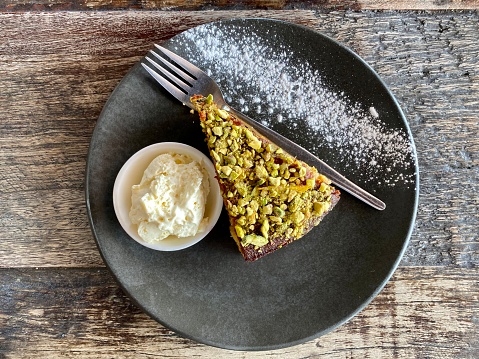 Horizontal high angle closeup photo of a slice of delicious Orange and Almond cake with chopped pistachios on top and a bowl of whipped cream, on a ceramic plate, on a rustic wooden table in a cafe