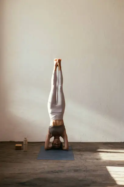 A portrait of a young Caucasian woman doing a headstand practicing yoga on her mat indoors
