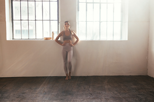 A wide angle horizontal portrait of a young blonde Caucasian woman standing in a gym in her sportswear barefoot