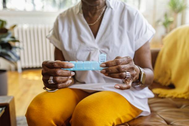 Woman taking daily dosage of medicines Senior black woman sits on the couch at home and takes medications from a daily pill organizer. Cropped shot does not show the woman's face. curing stock pictures, royalty-free photos & images