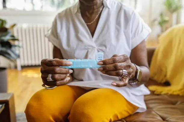 Senior black woman sits on the couch at home and takes medications from a daily pill organizer. Cropped shot does not show the woman's face.