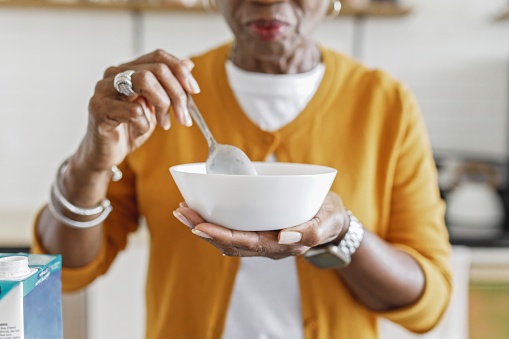 Close up shot of a senior African American woman holding a bowl of cereal for breakfast while standing in the kitchen on her home. Selective focus on the bowl and spoon in her hands.