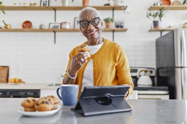 retrato de uma mulher idosa usando um tablet enquanto tomando café da manhã na cozinha - muffin freedom breakfast photography - fotografias e filmes do acervo