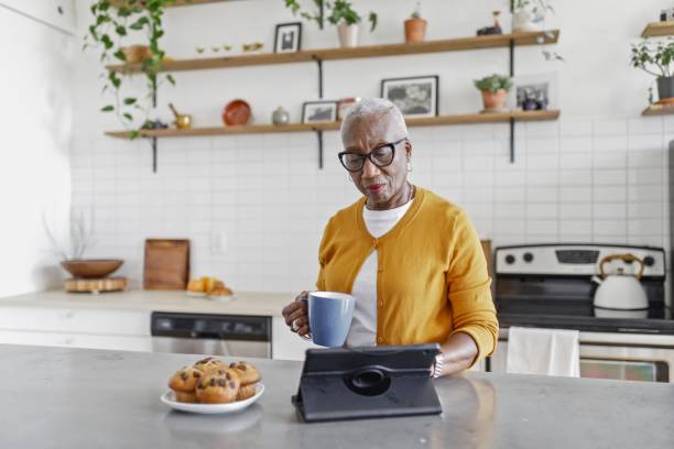 donna anziana lavora sul suo tablet mentre fa colazione nella sua cucina - muffin freedom breakfast photography foto e immagini stock
