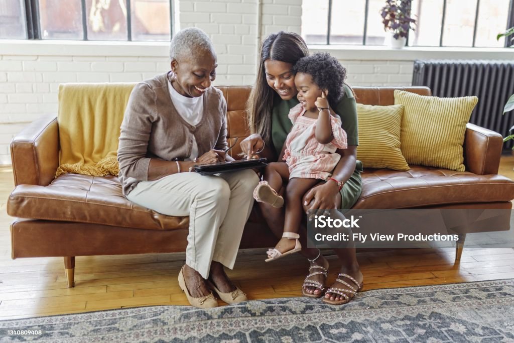 Multi-generation family bonding at home Adorable mixed race toddler girl sits on her Indian mother's lap. The child's grandmother is visiting. The black senior woman is showing her daughter-in-law and granddaughter something on a tablet computer. The multi-generation family is enjoying a relaxing day together at home. Multi-Generation Family Stock Photo