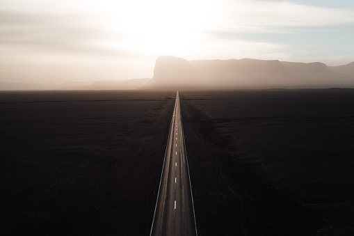 Endless straight road into the sun between black volcanic gravel towards sunset over icelandic mountain range. Aerial drone point of view along the country highway. Toned Image Colors. Southern Iceland, Golden Circle Route, Iceland, Northern Europe.