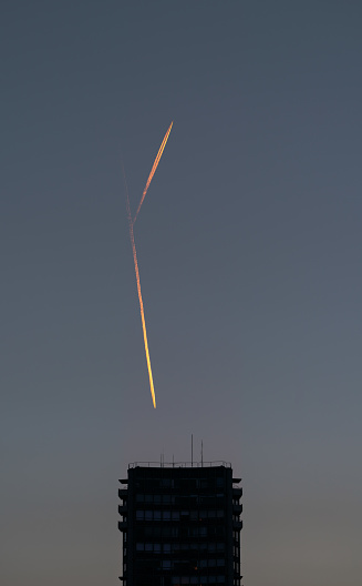 A closeup silhouette of a highrise building with two jet planes diverging with contrail vapor trails left behind and glowing in the sunset light on a blue sky backdrop.