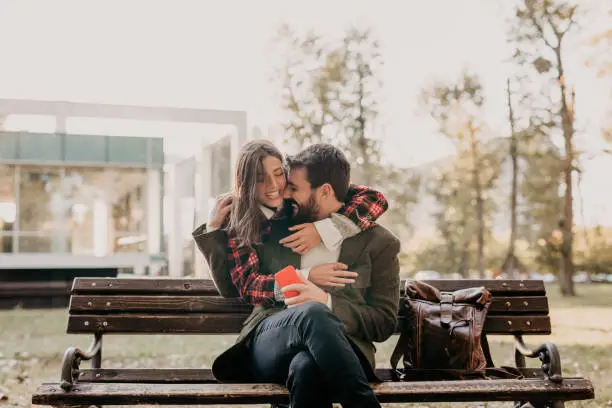 Spontaneous photo of a handsome bearded young adult man sitting on a wooden bench, outdoors in a park, holding the phone, with a cool backpack next to him and his beautiful partner is came to surprise and hug him from behind. Gentle kissing. Both stylishly dressed, in retro coats, in layers
