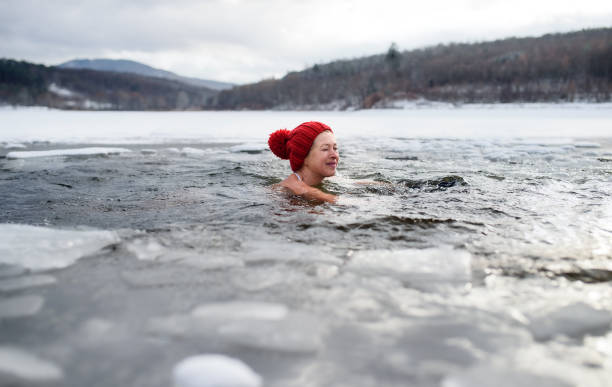 Happy active senior woman in hat diving outdoors in water in winter, cold therapy concept. Copy space. A happy active senior woman in hat diving outdoors in water in winter, cold therapy concept. Copy space. woman cold water swimming stock pictures, royalty-free photos & images
