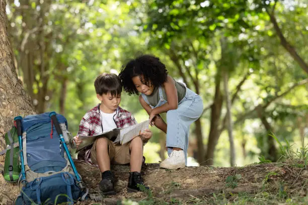 Photo of African American ethnicity boy and girl children wearing plaid shirt and backpack sitting at tree base talking and looking down on the map travel on hand in the park background hiking adventure summer holiday concept