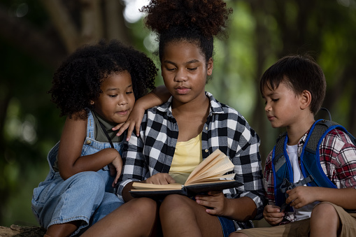 Sister black skin African American ethnicity sitting at on tree base nature reading to her younger siblings a book about adventure stories listen in natural forest