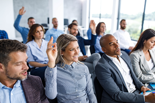 Large group of business people attending an education event in a board room. Focus is on happy woman raising her hand to ask a question.