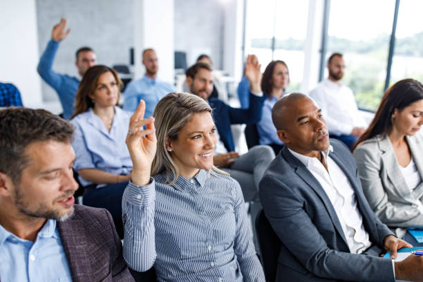 Engaging seminar in board room! Large group of business people attending an education event in a board room. Focus is on happy woman raising her hand to ask a question. education event stock pictures, royalty-free photos & images