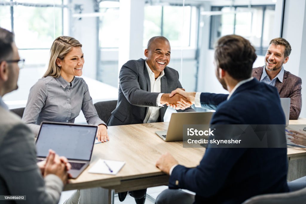 Happy businessmen shaking hands on a meeting in the office. Happy male entrepreneurs came to an agreement during a meeting with their colleagues in the office. Focus is on black man. Handshake Stock Photo