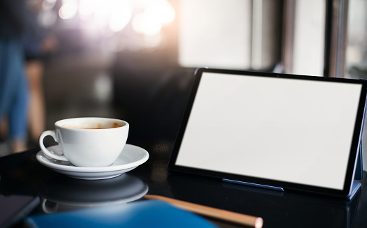 Focus on white coffe cup with blank screen of digital tablet on desk in cafe