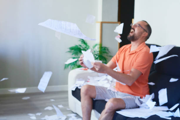 un jeune homme hipster avec une barbe assis sur le canapé à la maison jetant des papiers en l’air tout en riant aux éclats à cause du stress - paper document flying throwing photos et images de collection