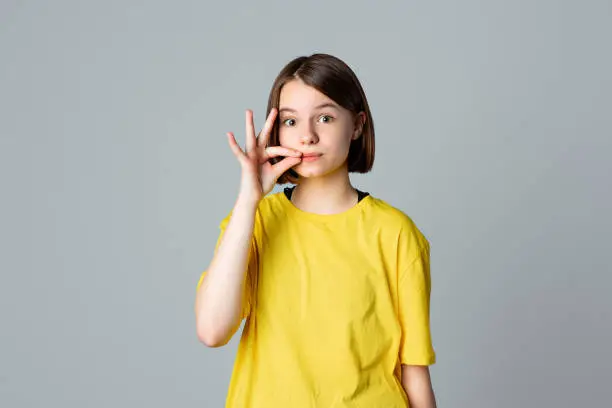 Portrait of a pretty teen girl showing zip gesture as if shutting mouth on key, standing over light grey background. You must be silent. Ban. taboo