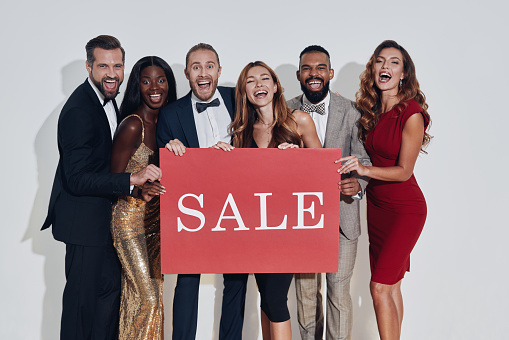 Group of beautiful people in formalwear holding sale banner and smiling while standing against gray background