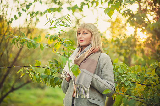 A blonde woman with a short haircut walks through the forest in a gray wool coat and a plaid scarf. Walking in nature in autumn forest