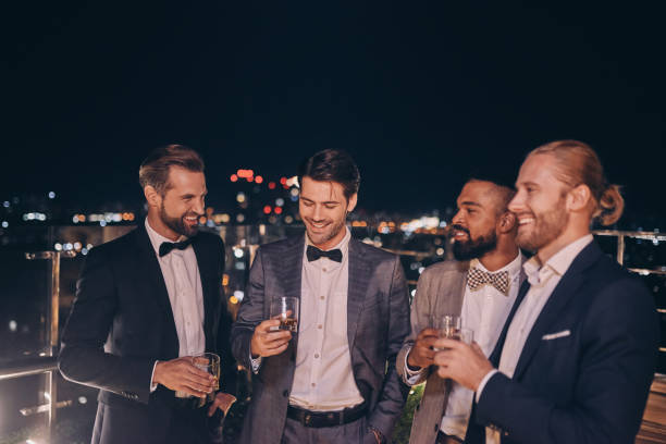 Group of handsome young men in suits and bowties drinking whiskey and smiling Group of handsome young men in suits and bowties drinking whiskey and smiling while spending time on outdoor party dressing up stock pictures, royalty-free photos & images