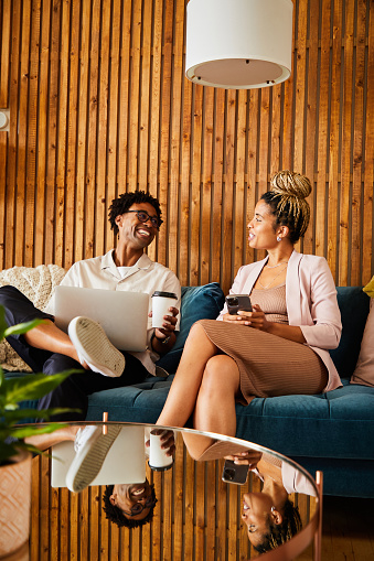Two laughing young African American businesspeople talking while using different devices while sitting on an office sofa during a break