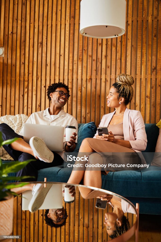 Laughing young businesspeople using different devices during an office break Two laughing young African American businesspeople talking while using different devices while sitting on an office sofa during a break Office Stock Photo