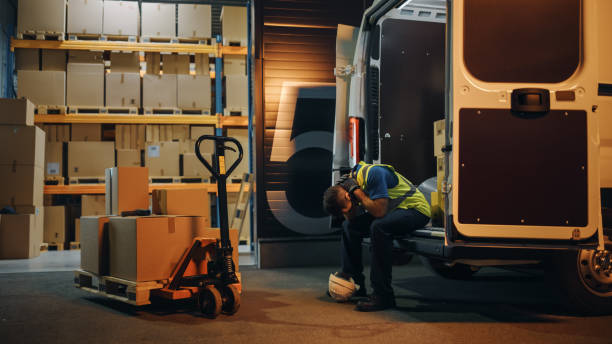 latin male worker wearing hard hat loads cardboard boxes into delivery truck, rests. online orders, e-commerce goods, food, medicine. tired overworked frontline hero. dramatic shot - logistical imagens e fotografias de stock