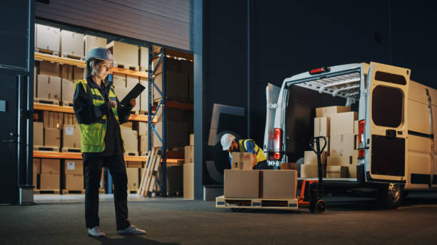 istock Portrait of Beautiful White Woman Worker Using Tablet Computer in Warehouse full Cardboard Boxes. In Background Professional Loading Cardboard boxes,  e-Commerce Online Orders into Truck. 1340887674