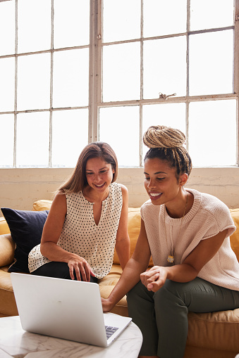 Two diverse young businesswomen sitting on an office sofa and smiling while working together on a laptop